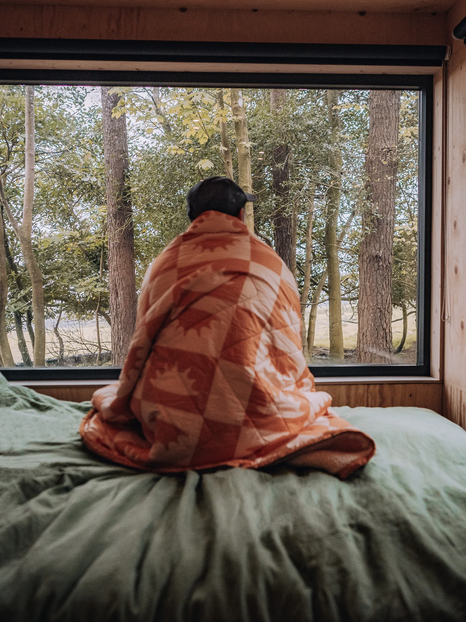 Man sitting on the bed wrapped in a blanket looking out at the woodland from the Unplugged Mabel cabin