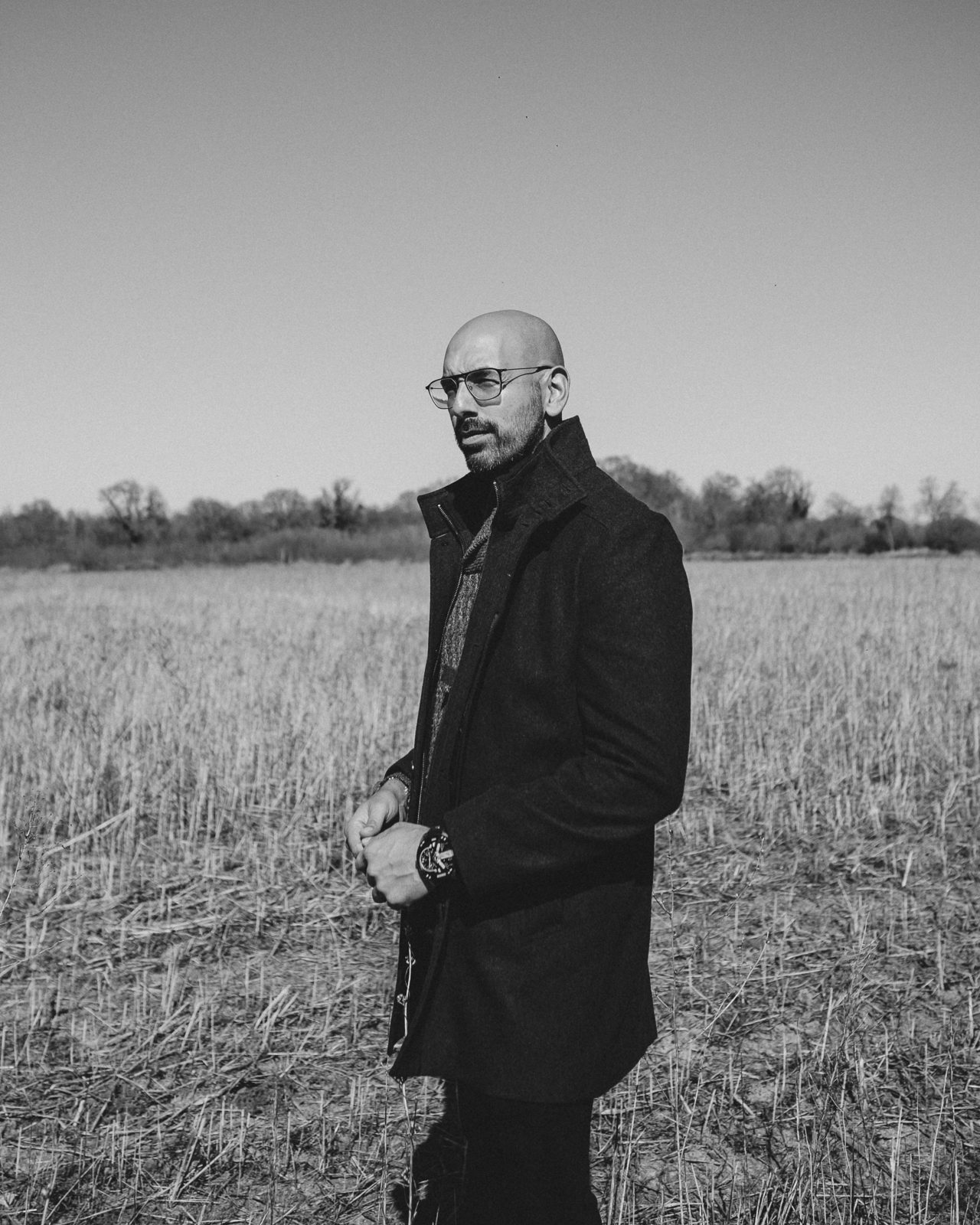 Man in a dark jacket and glasses stands in. grass field.