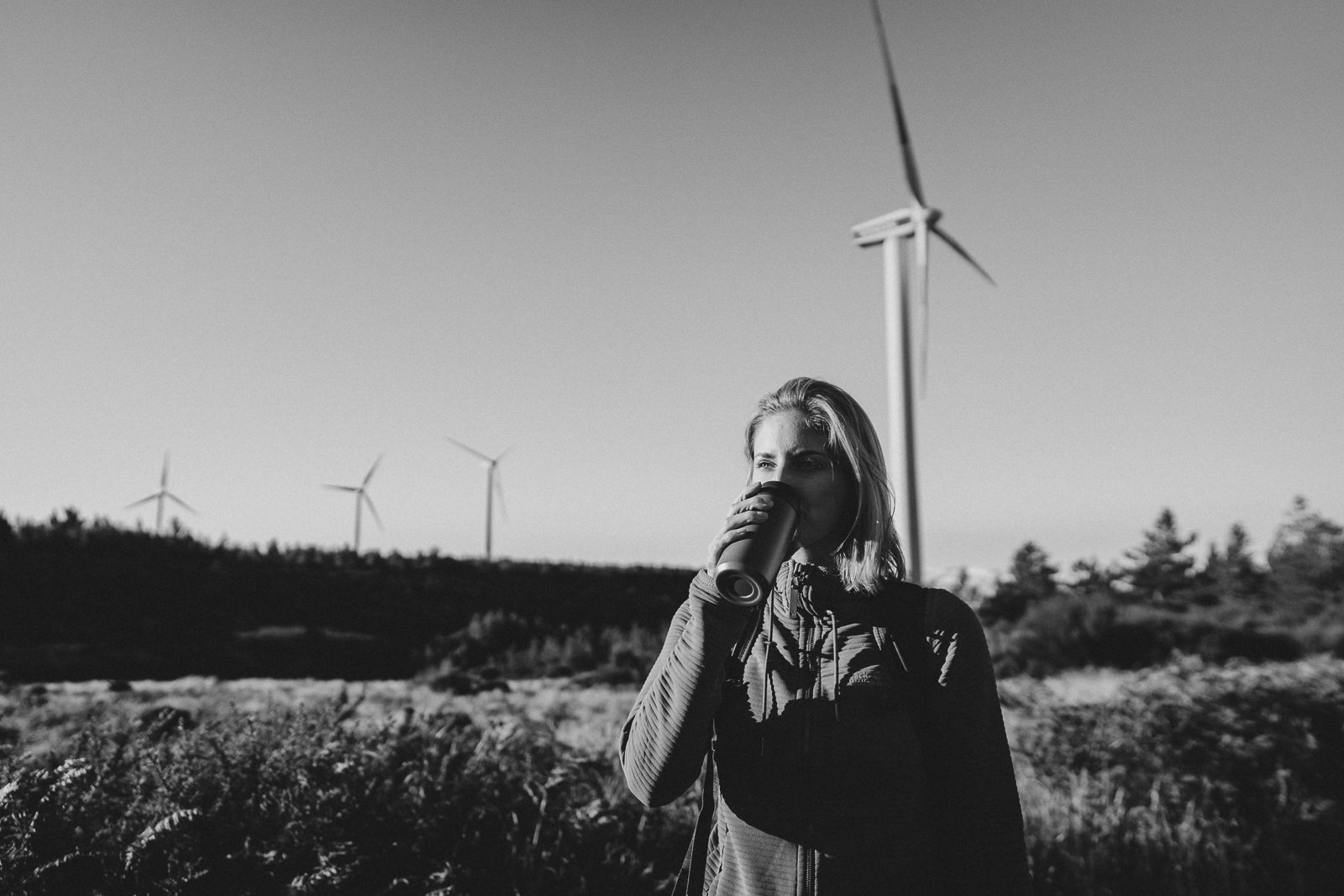 Woman drinking a coffee stands in a field of wind turbines.