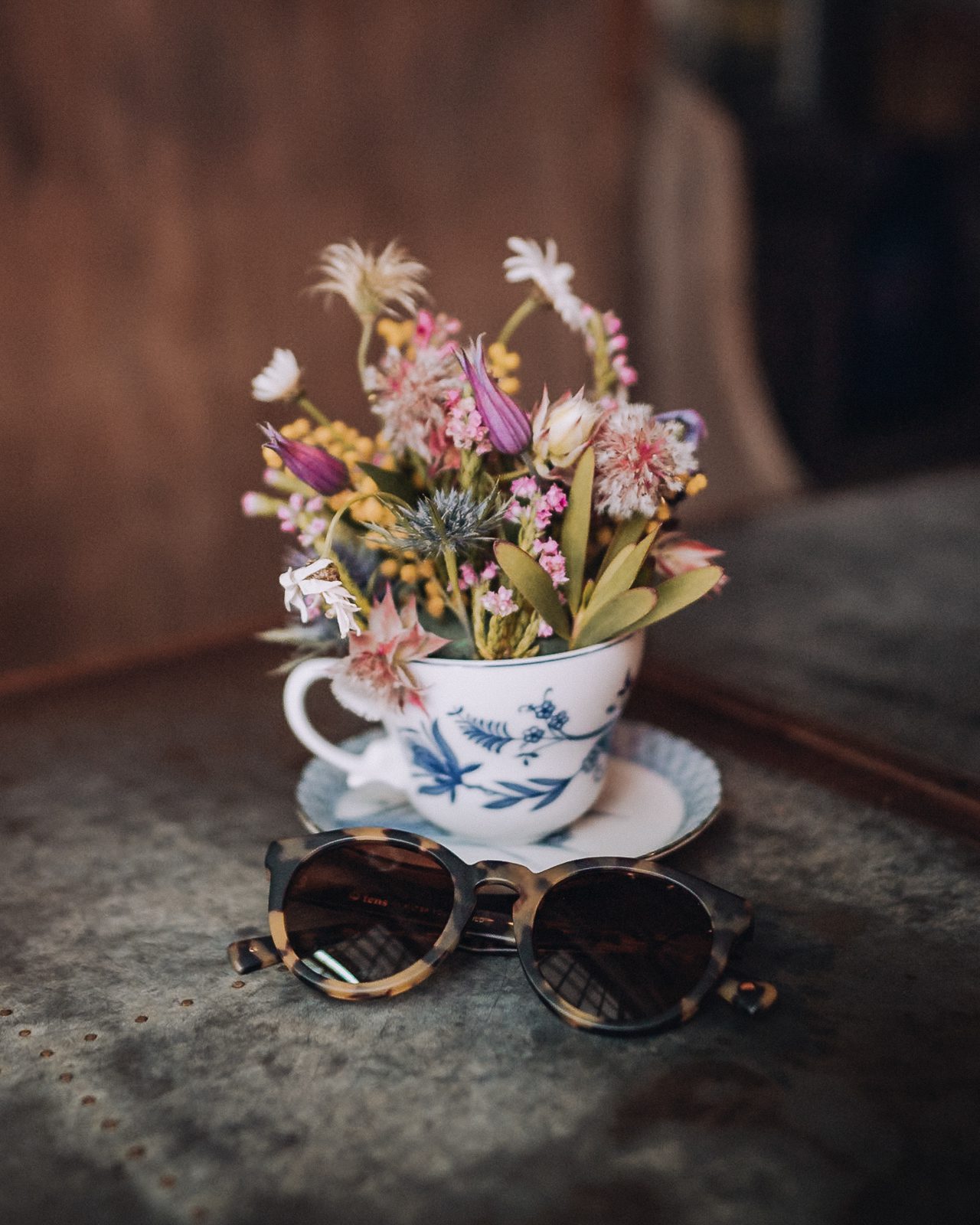 A pair of tortoiseshell sunglasses next to a flower filled teacup.