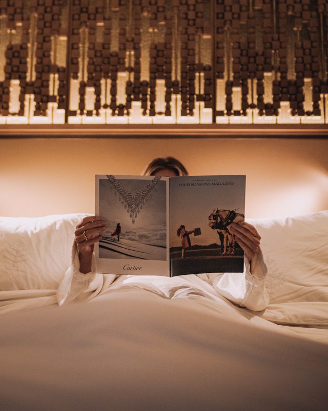 Woman reads a magazine while sitting in luxurious hotel bed.
