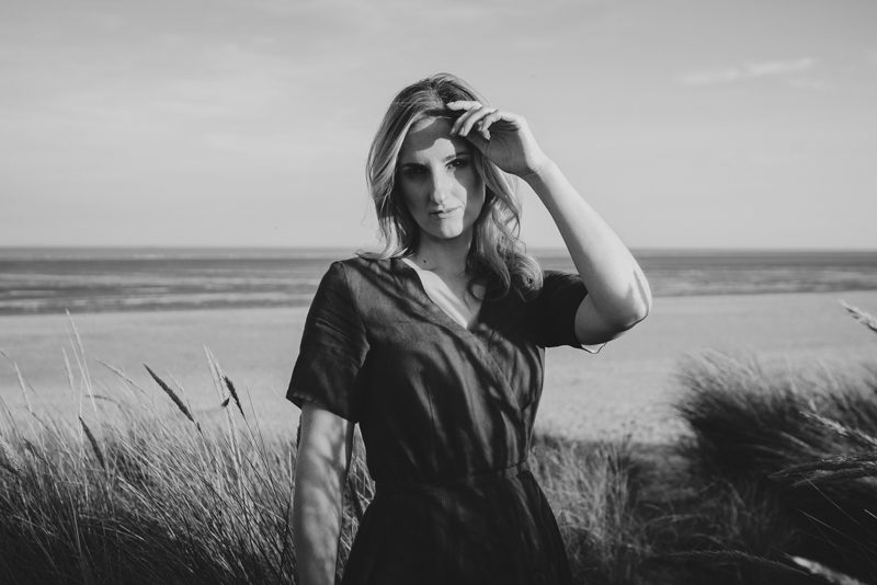 Woman stands amongst dunes on a stretch of sandy beach.