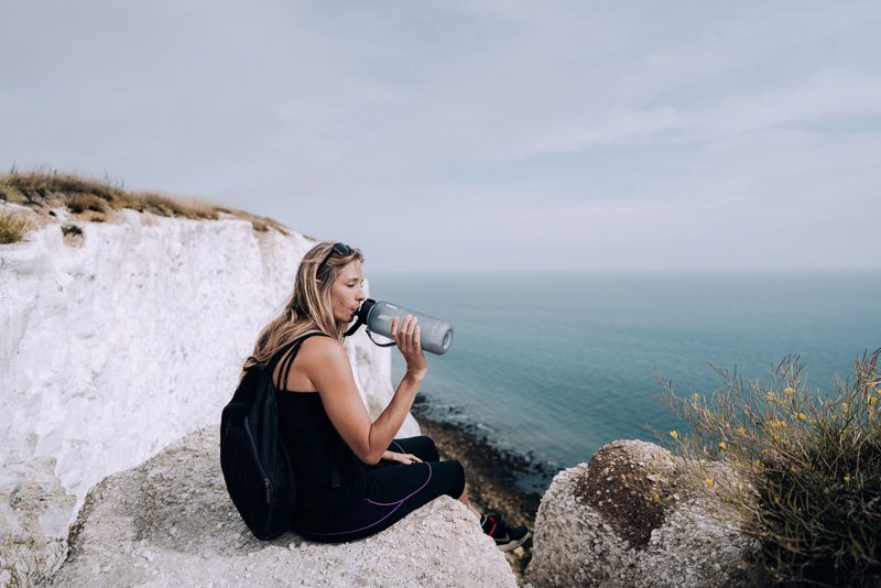 Woman sits on white chalk cliffs while drinking from sports bottle.